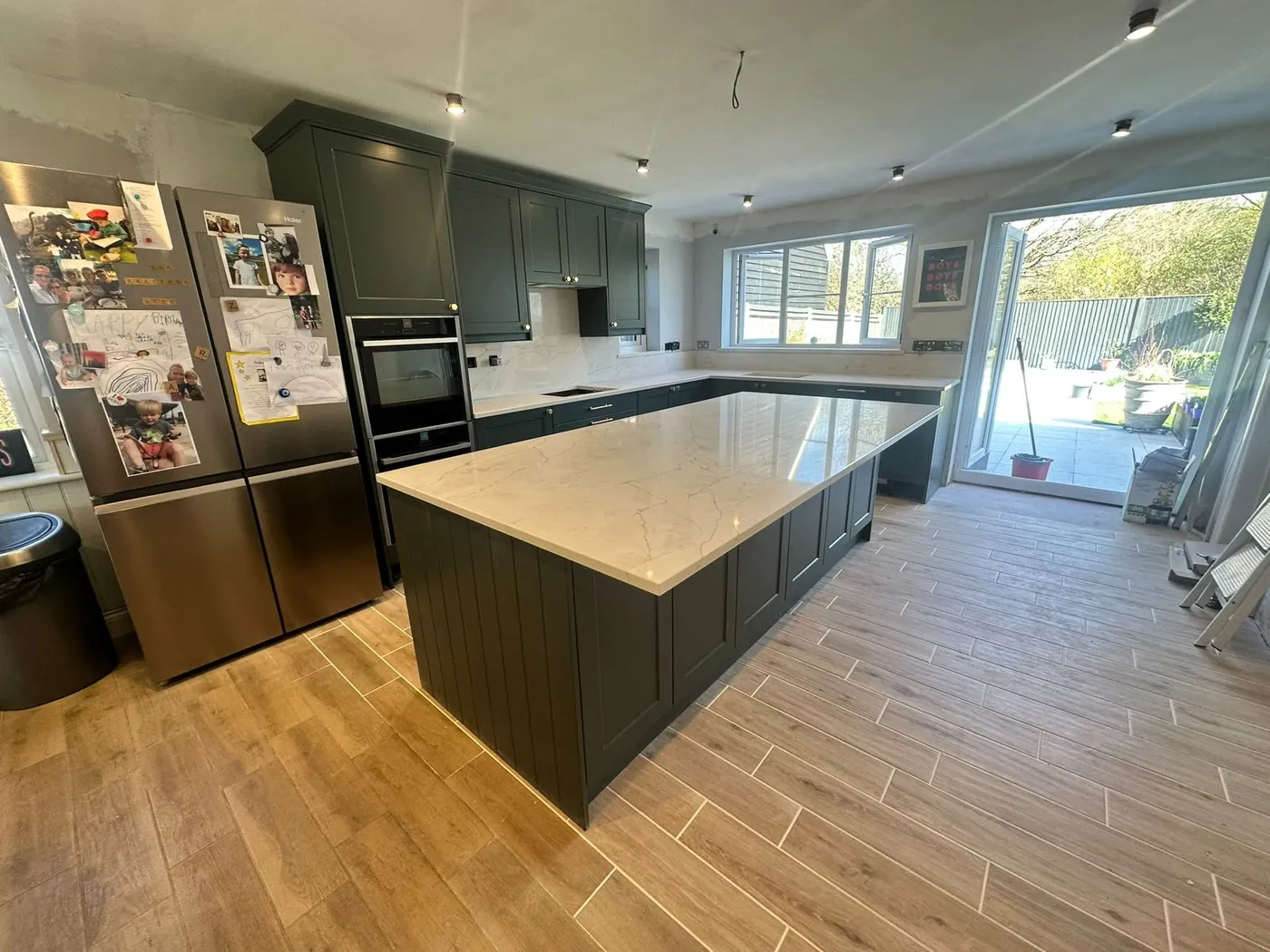 Open-plan dark green shaker kitchen with a marble-effect island, tall oven housing and garden doors
